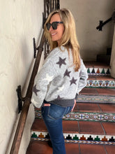 A woman wearing a light gray knit sweater with dark gray star patterns and dark sunglasses, standing on a tiled staircase indoors.