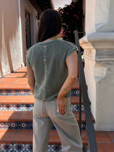 A woman viewed from behind wearing a ribbed sage green short-sleeve top with buttons down the back and light beige pants, standing on terracotta tiled steps.