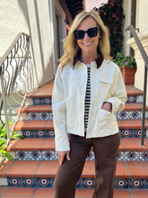 A woman wearing a cream-colored canvas utility jacket with dark brown collar and pocket trim, styled over a black and white striped shirt and brown pants, standing on outdoor tiled steps.