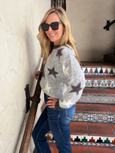 A woman wearing a gray fuzzy sweater with star and crescent moon patterns, blue jeans, and black sunglasses, posing on a tiled staircase.