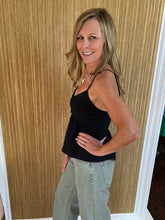 Woman wearing a black top and light olive jeans standing against a wooden paneled wall.