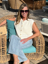 Woman sitting on a wicker chair with a teal cushion, wearing sunglasses and a white sleeveless top.