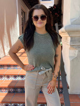 A woman wearing a ribbed sage green short-sleeve top and beige pants stands on tiled outdoor steps. She is wearing round sunglasses and has visible tattoos on her arms.