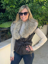 A woman wearing a dark brown quilted wrap vest with a large gray faux fur collar, a cream ribbed turtleneck, and dark jeans, posing outdoors in front of green foliage.