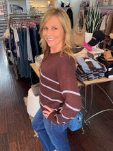A smiling blonde woman wearing a brown and white horizontally striped knit sweater and blue jeans, standing inside a clothing boutique with racks of garments and tables visible in the background.