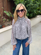 A woman wearing a gray and white abstract patterned long-sleeve blouse with a high gathered neckline, paired with blue jeans and black sunglasses, standing outdoors on a concrete walkway with green plants and a wooden fence in the background.