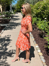 Woman in an orange floral dress standing on a garden path with greenery around.