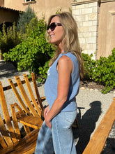 Woman in a blue sleeveless top and jeans standing outdoors near wooden chairs.