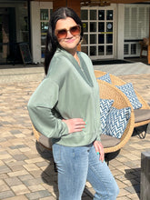 Woman wearing a green sweatshirt and sunglasses standing outdoors near a wicker chair with cushions.