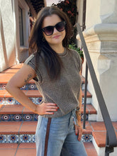 A woman wearing a brown and beige vertically striped knit top, light blue jeans, and dark sunglasses, posing on outdoor terracotta steps with a stone wall background.