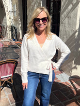 A woman wearing a cream-colored wrap cardigan and blue jeans, standing outdoors on a brick patio in front of a stucco wall.