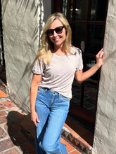 A woman with blonde hair wearing a light beige short-sleeve t-shirt and blue jeans, posing outdoors against a textured stucco wall and glass door.