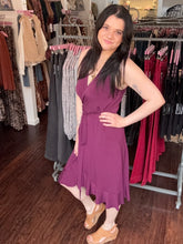 A woman modeling a sleeveless wrap-style midi dress in a rich plum purple color, standing inside a clothing boutique with racks of garments in the background.