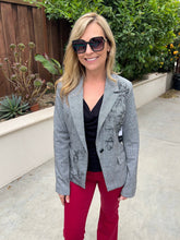 A woman wearing a light gray textured blazer with black floral embroidery, a black top, and bright magenta pants, standing outdoors in front of a wooden fence and green plants.
