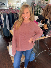 A woman with blonde hair stands in a clothing boutique wearing a dusty rose pink ribbed knit sweater and blue jeans. Clothing racks, a table with shoes, and store fixtures are visible in the background.