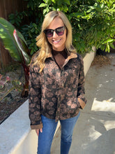 A woman wearing a brown and black floral pattern barn jacket , paired with blue jeans and sunglasses, standing outdoors on a concrete walkway with green foliage in the background.
