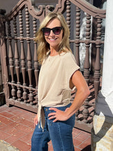 A woman wearing a beige short-sleeve tie-front top and blue jeans stands outdoors in front of a decorative brown metal railing.