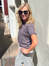 A woman wearing a short-sleeve crewneck t-shirt in a muted purple-gray color and light gray patterned sweatpants, standing outdoors against a textured white wall.