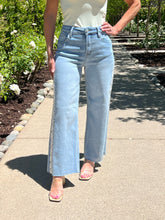 Person wearing light blue jeans standing on a sidewalk with greenery in the background