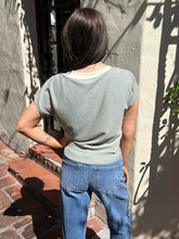 A woman wearing a light sage green short-sleeve knit top and blue jeans stands outdoors against a stucco wall with shadows.