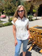 Woman in a white shirt and blue jeans standing outdoors with trees and a building in the background.