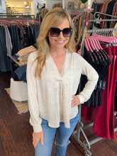 A woman wearing a cream-colored long-sleeve blouse with a subtle leopard print pattern, paired with blue jeans and dark sunglasses, standing inside a clothing boutique.
