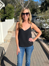 Woman wearing a black tank top and blue jeans with trees in the background.