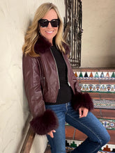A woman wearing a brown faux-leather moto jacket with dark brown fur trim on the collar and cuffs, styled with a black top and blue jeans, leaning against a textured wall on a tiled staircase.