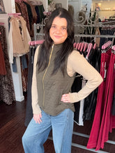 A woman posing in a boutique wearing an olive green quilted sleeveless vest over a beige long-sleeve knit top and blue jeans, with clothing racks visible in the background.
