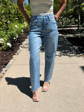 Person wearing light blue jeans standing on a sidewalk with greenery in the background