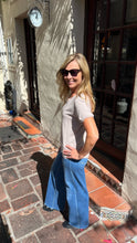A woman wearing a light beige short-sleeve t-shirt and wide-leg blue denim jeans stands on a brick sidewalk outside a building with a stucco wall and dark wood doors.