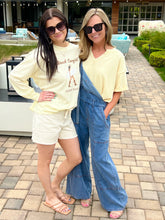 Two women standing on a patio, one in a white 'Beach Cowgirl' shirt and shorts, the other in a yellow top and blue overalls.