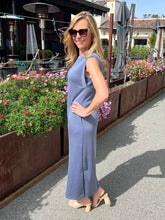 Woman in a blue sleeveless dress standing outdoors with flowers and outdoor furniture in the background.