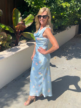 Woman in a light blue floral dress standing outdoors with greenery in the background
