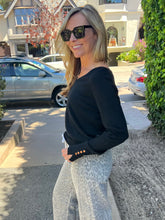 A woman wearing a black long-sleeve top with gold button details on the cuff and light gray leopard-print pants, standing outdoors on a residential street.