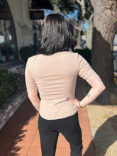 Person wearing a khaki tee and black pants standing outdoors with trees and a building in the background.