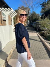 Woman wearing a navy blue top and white pants standing on a wooden deck with trees and a house in the background.
