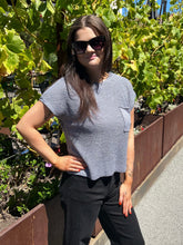 A woman wearing a gray ribbed short-sleeve crop top with a chest pocket and black pants, standing outdoors in front of green leafy plants.