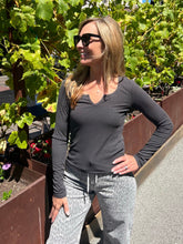 A woman wearing a dark charcoal gray long-sleeve V-neck top and gray leopard-print pants, standing outdoors in front of green grapevines.