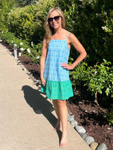 Woman wearing a blue and green dress standing on a sidewalk with greenery in the background