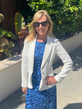 Woman wearing a white blazer over a blue dress with sunglasses, standing outdoors.
