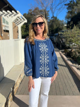 Woman wearing a blue embroidered blouse and white pants standing on a wooden deck.