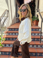 A woman wearing a cream long-sleeve button-up shirt and brown wide-leg pants, standing on tiled outdoor stairs.