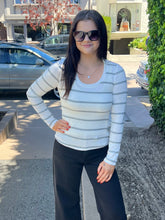 A woman wearing a white long-sleeve scoop-neck knit top with thin gray horizontal stripes, paired with black wide-leg pants, standing outdoors on a sidewalk.