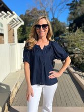 Woman wearing a navy blue top and white pants standing on a wooden deck with trees and a house in the background.