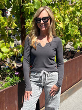 A woman wearing a dark gray long-sleeve V-neck top and light gray patterned sweatpants, standing outdoors in front of green plants and a brown planter box.