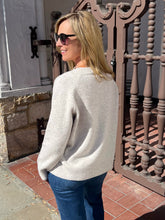 A woman wearing a light beige knit sweater and blue jeans stands outdoors in front of a brown metal gate and stone wall, shown from the back.