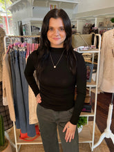 A woman with long dark hair stands in a clothing boutique wearing a fitted black long-sleeve mock neck top and dark gray jeans. She is positioned in front of white clothing racks and shelves filled with garments.
