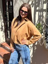 A woman wearing a tan suede zip-up jacket and blue jeans, standing outdoors in front of a stucco wall with shadows.
