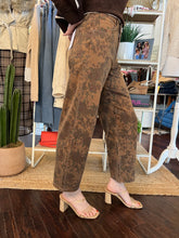 A person wearing brown wide-leg cropped pants with a mottled tan and medium brown abstract pattern, styled with beige block-heel sandals in a boutique setting.